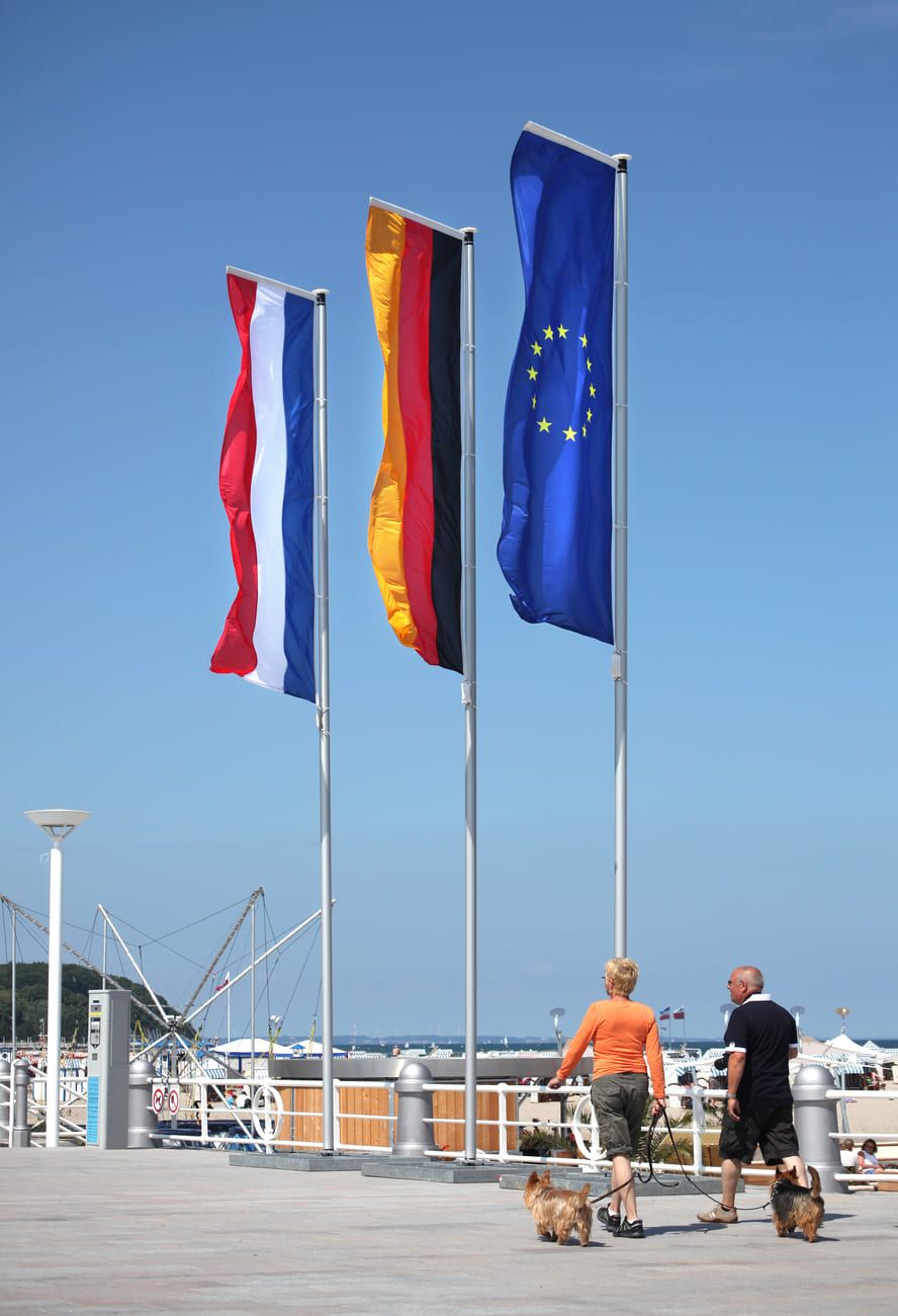 Eine deutsche, niederländische und europäische Fahne wehen an einem Fahnenmast am Strand im Wind mit Menschen davor