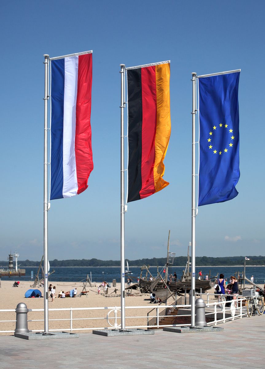Eine deutsche, niederländische und europäische Fahne wehen an einem Fahnenmast am Strand im Wind