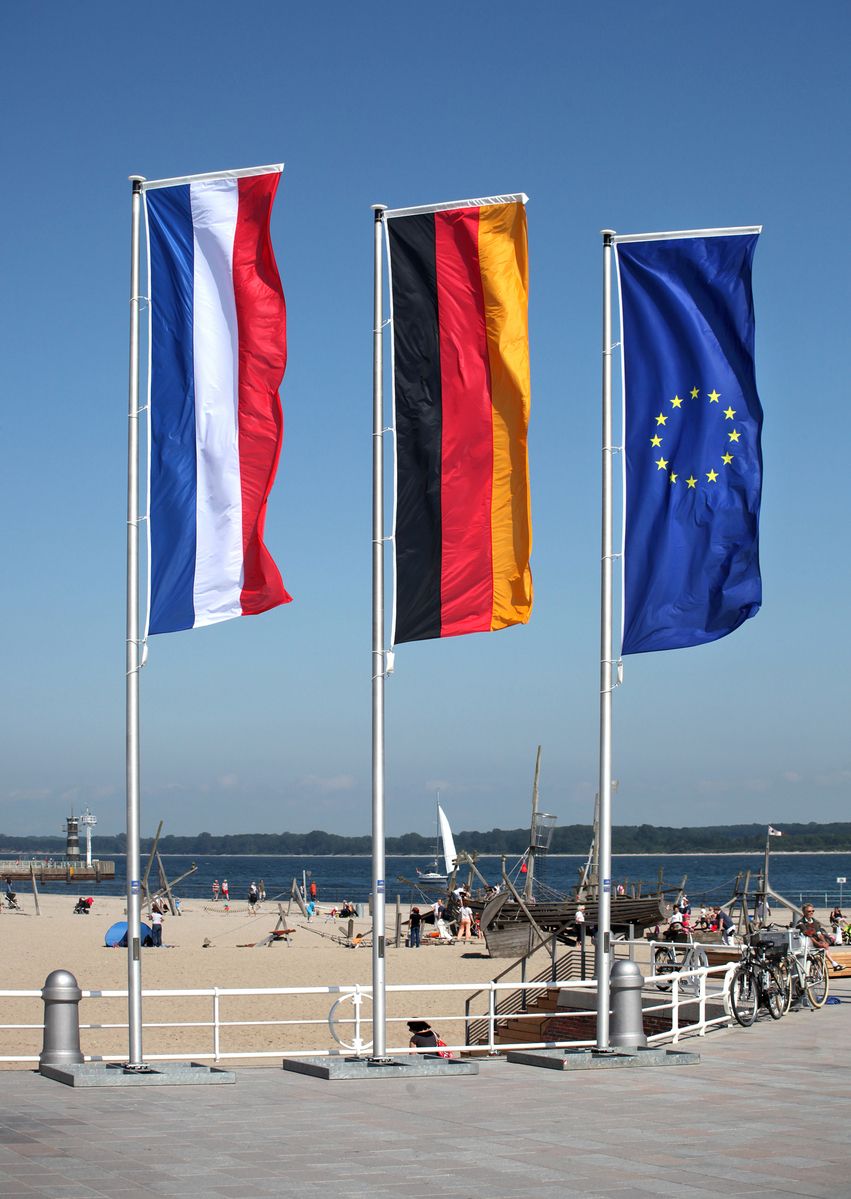 Eine deutsche, niederländische und europäische Fahne wehen an einem Fahnenmast am Strand im Wind
