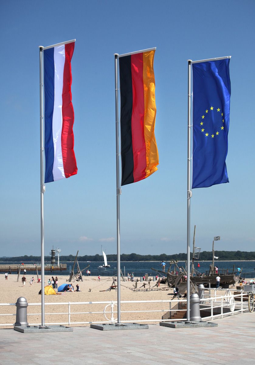 Eine deutsche, niederländische und europäische Fahne wehen an einem Fahnenmast am Strand im Wind