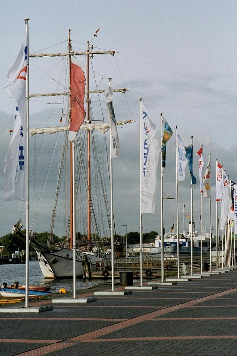 Mehrere Fahnenmäste stehen in einem Hafen vor einem alten Segelboot im Hintergrund