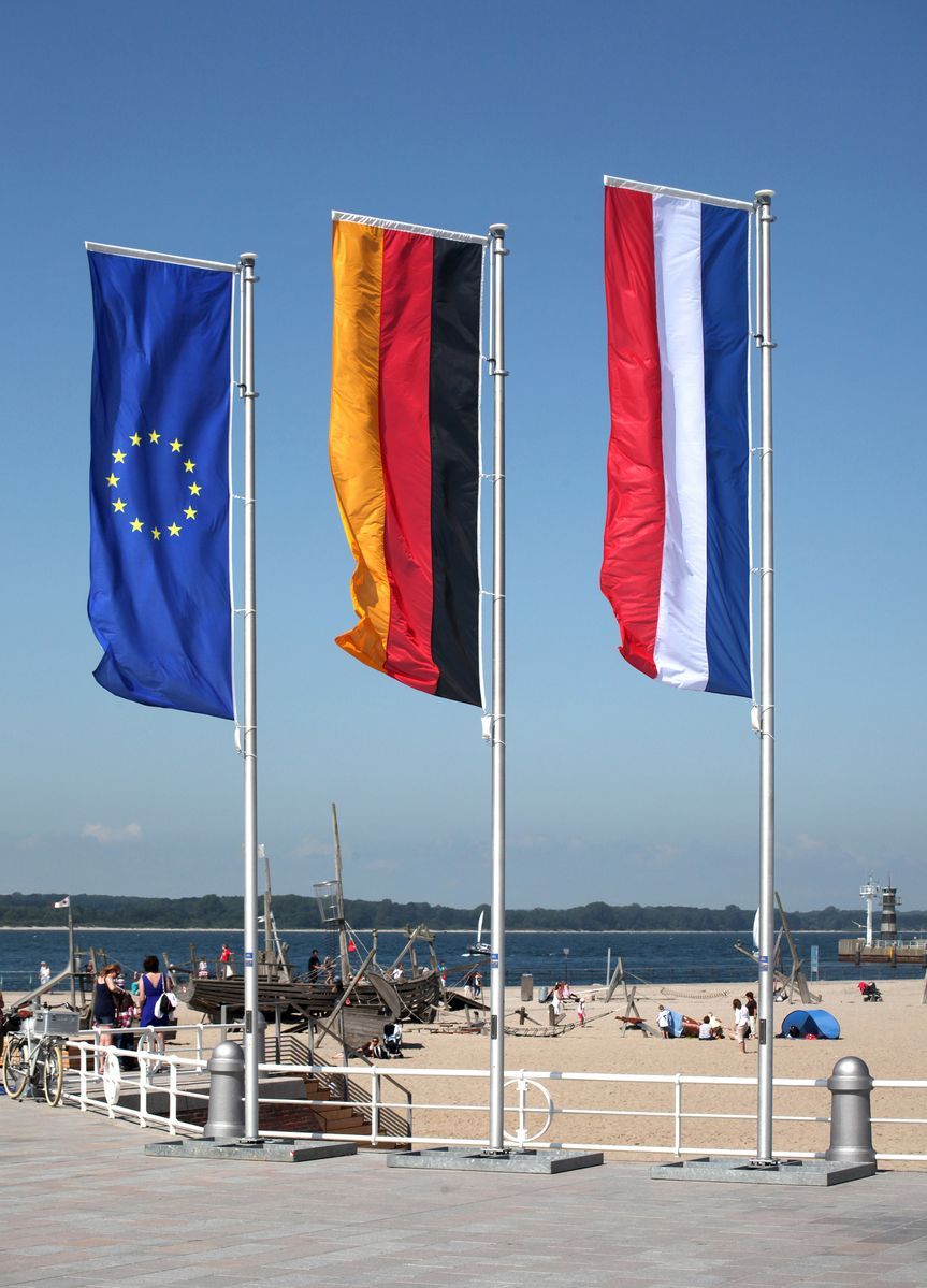 Eine deutsche, niederländische und europäische Fahne wehen an einem Fahnenmast am Strand im Wind