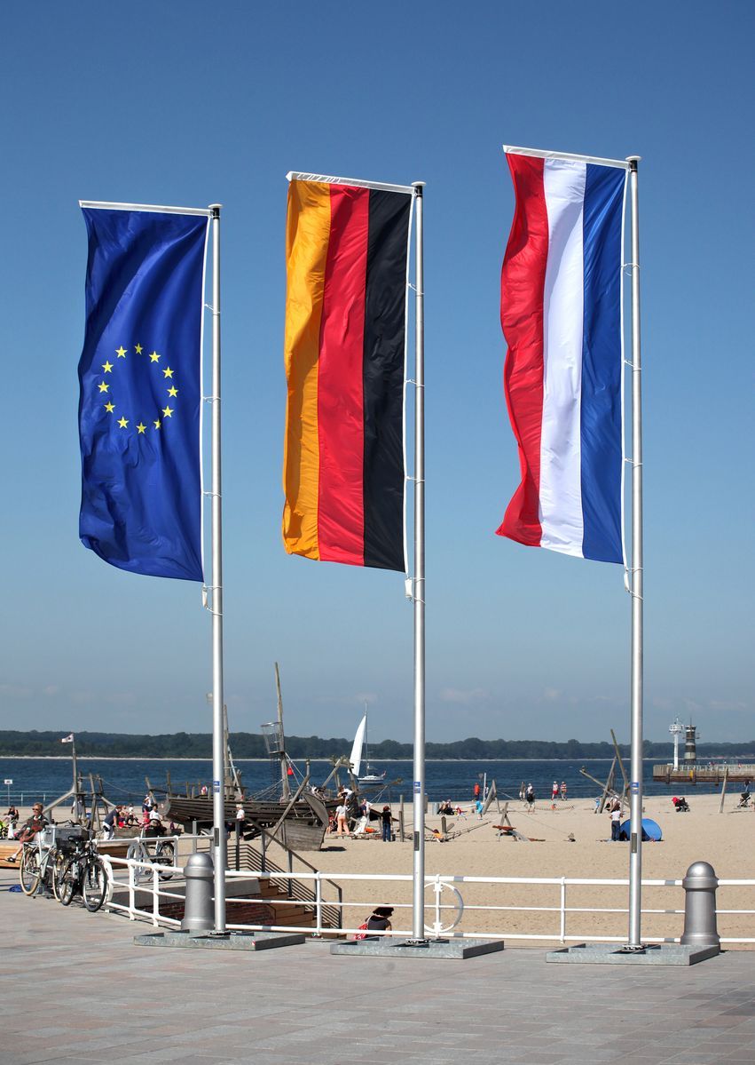 Eine deutsche, niederländische und europäische Fahne wehen an einem Fahnenmast am Strand im Wind